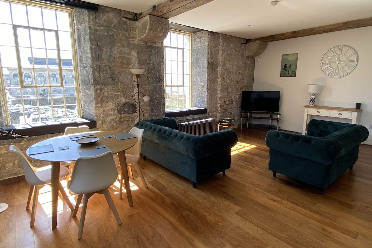 Un salon avec un mur en pierre, un canapé bleu, une table à manger avec des chaises, un radiateur et un téléviseur.