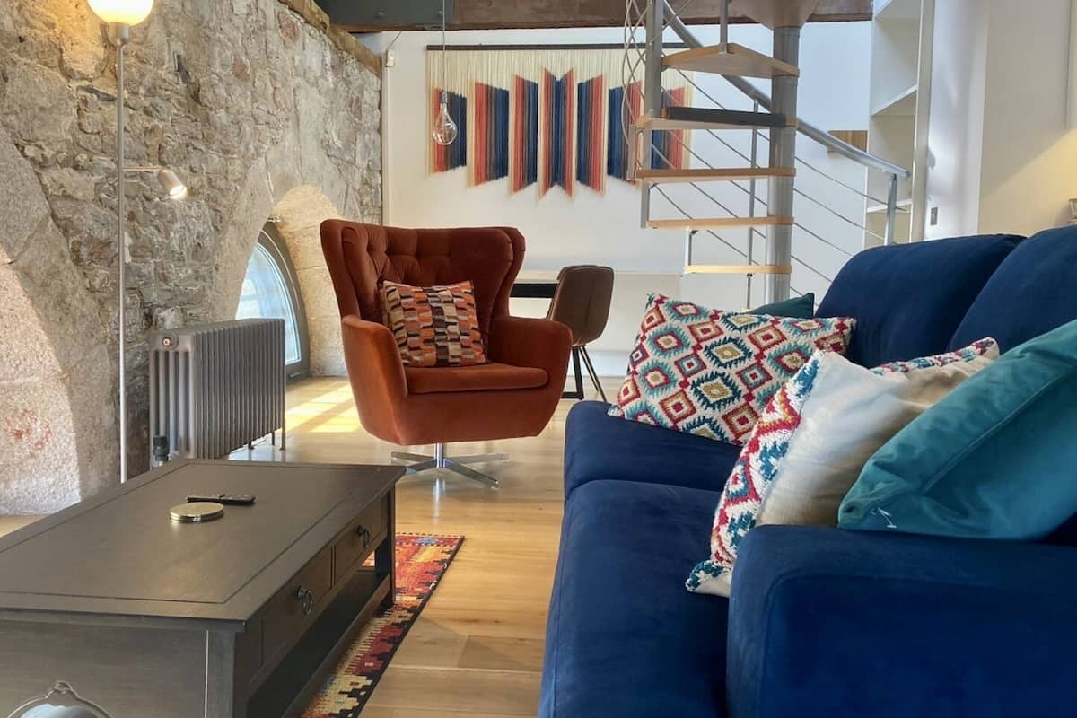 Un salon moderne avec un mur en pierre, un canapé bleu, un fauteuil orange et un escalier en bois.