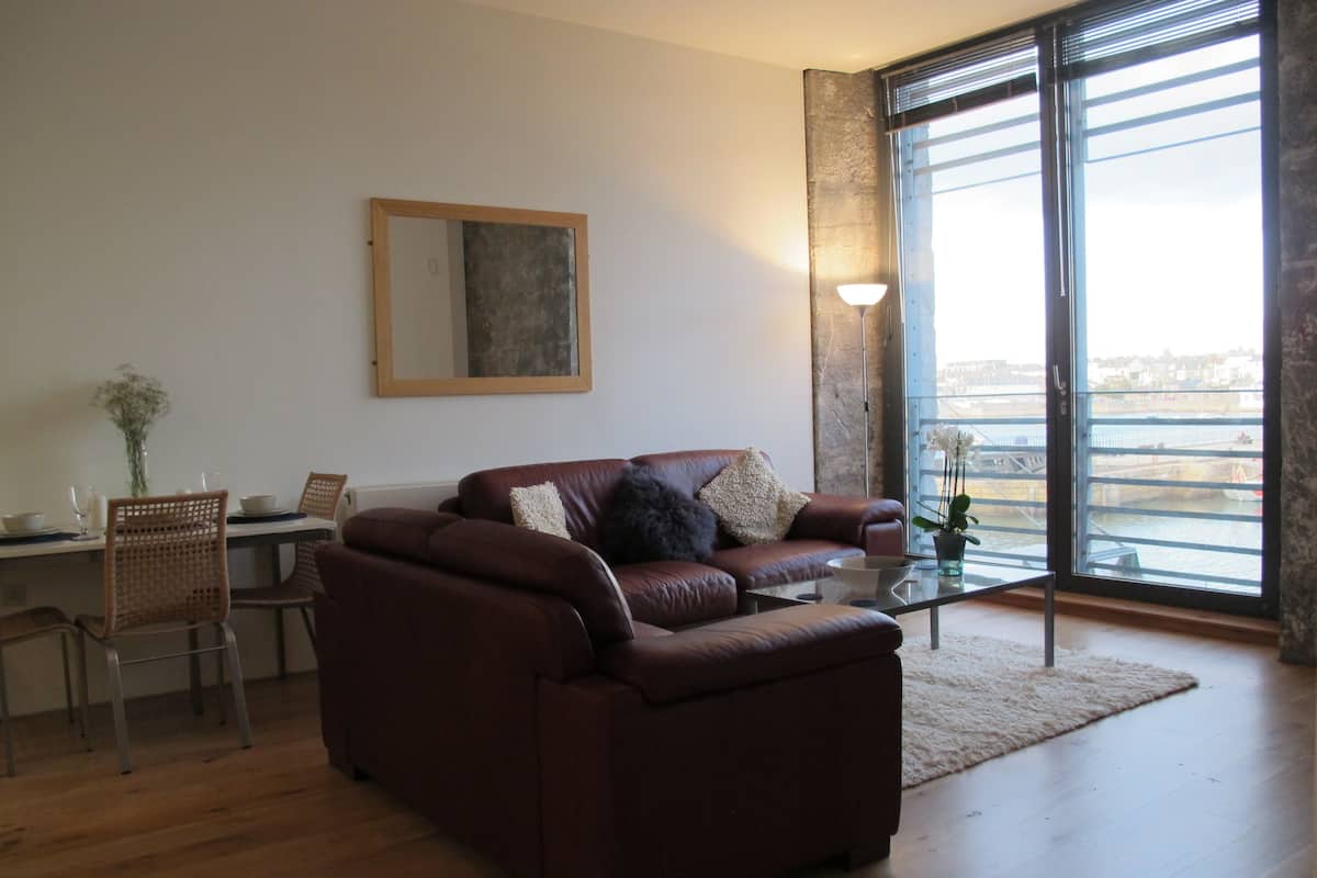 Un salon avec un canapé en cuir marron, une table à manger avec des chaises, une table basse en verre et une grande fenêtre donnant sur la ville.
