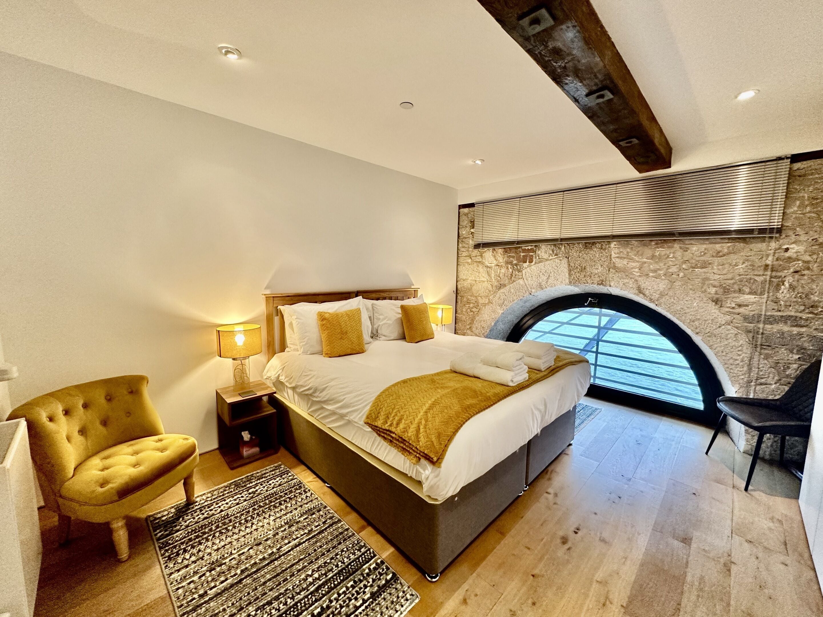A modern bedroom with a stone wall, a large bed, a yellow armchair, and a view through a window.