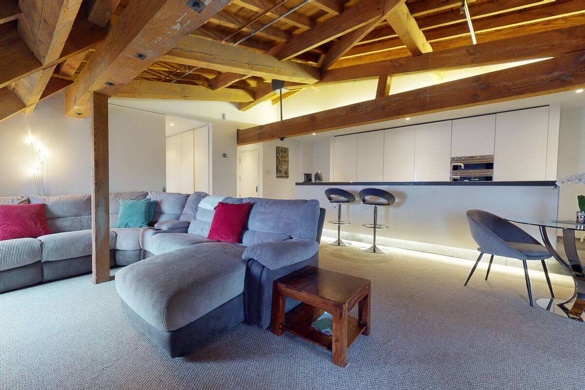 Un salon moderne avec un canapé gris, une table basse en bois et un coin cuisine doté de meubles blancs et d’un tabouret de bar.
