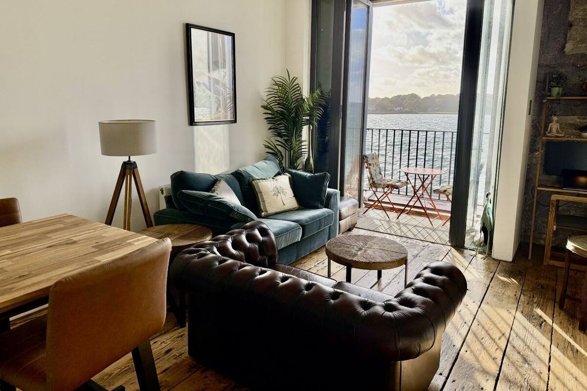 Un salon chaleureux avec un plancher en bois, un canapé en cuir, un divan bleu, une table à manger et un balcon offrant une vue sur la mer.