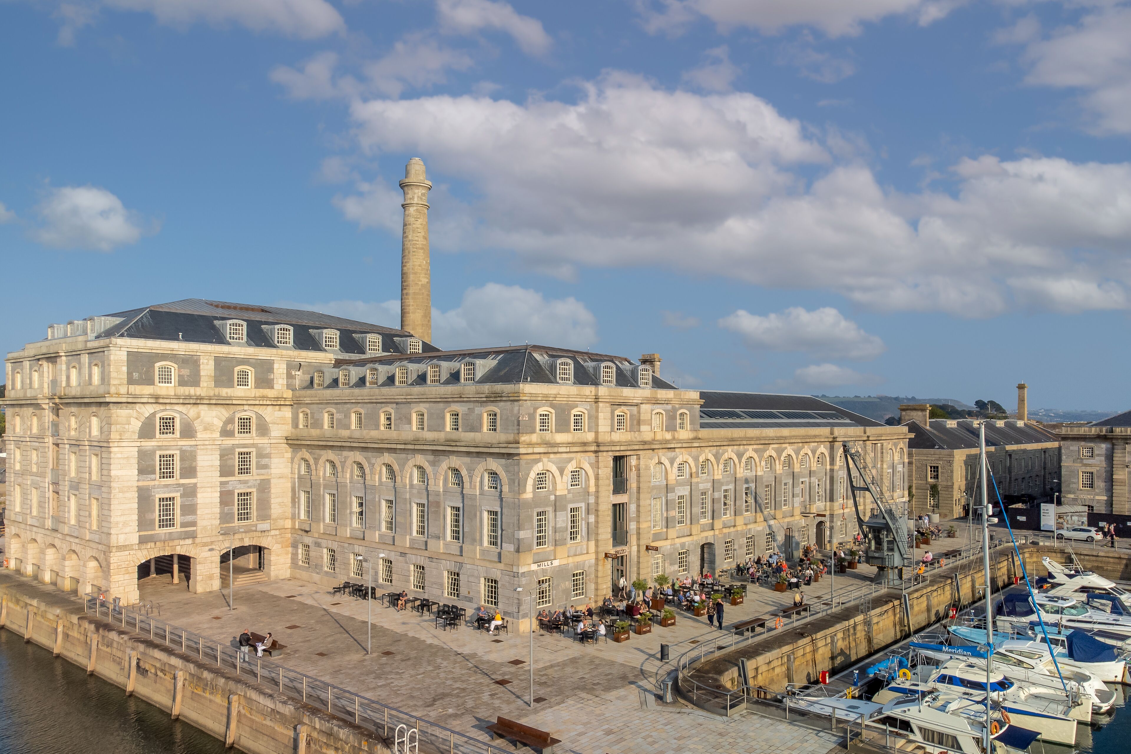 A historic building with a prominent tower, a waterfront promenade, and a marina with boats.