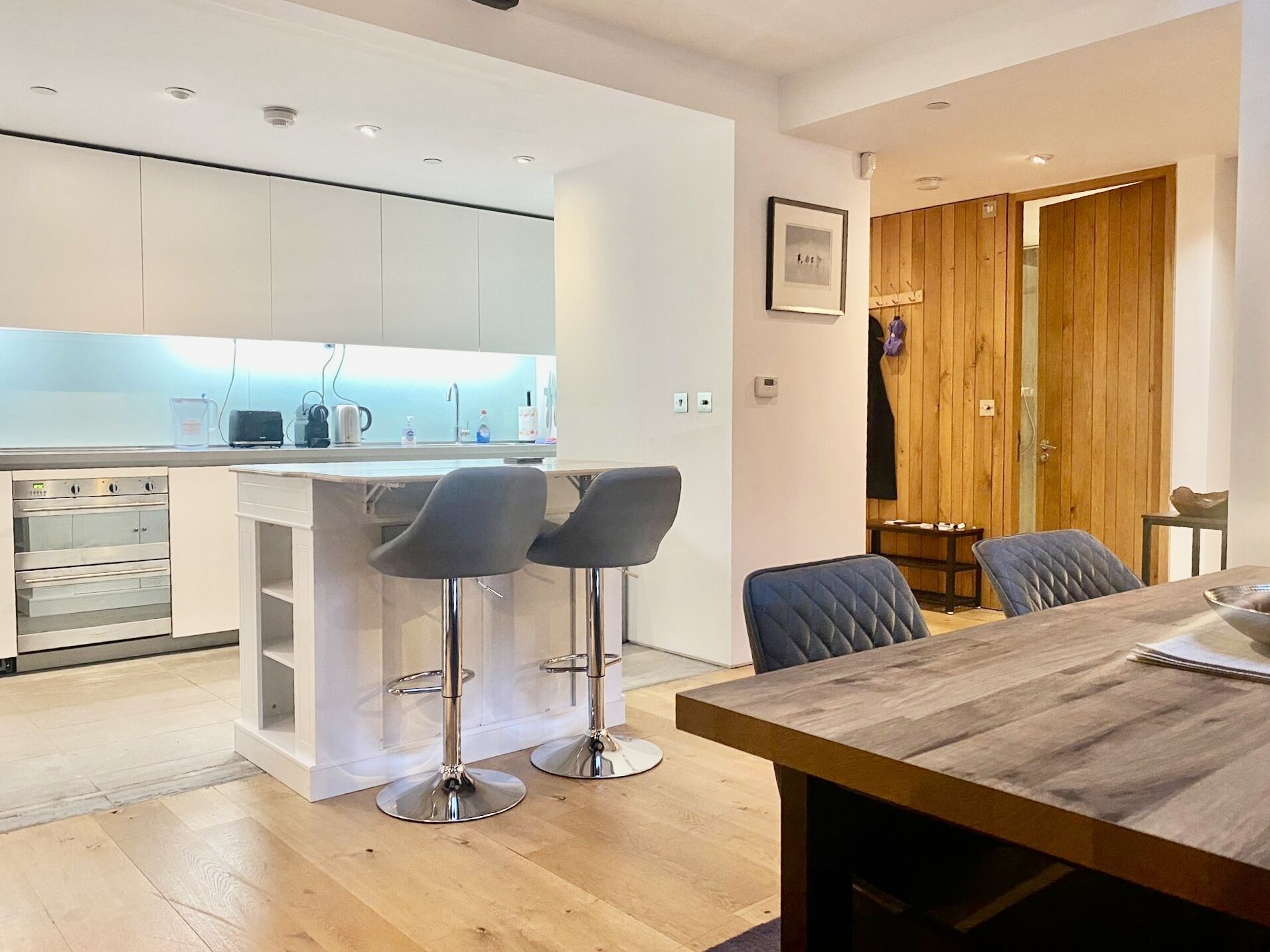 A modern kitchen with white cabinets, a built-in oven, and a bar area with two stools.