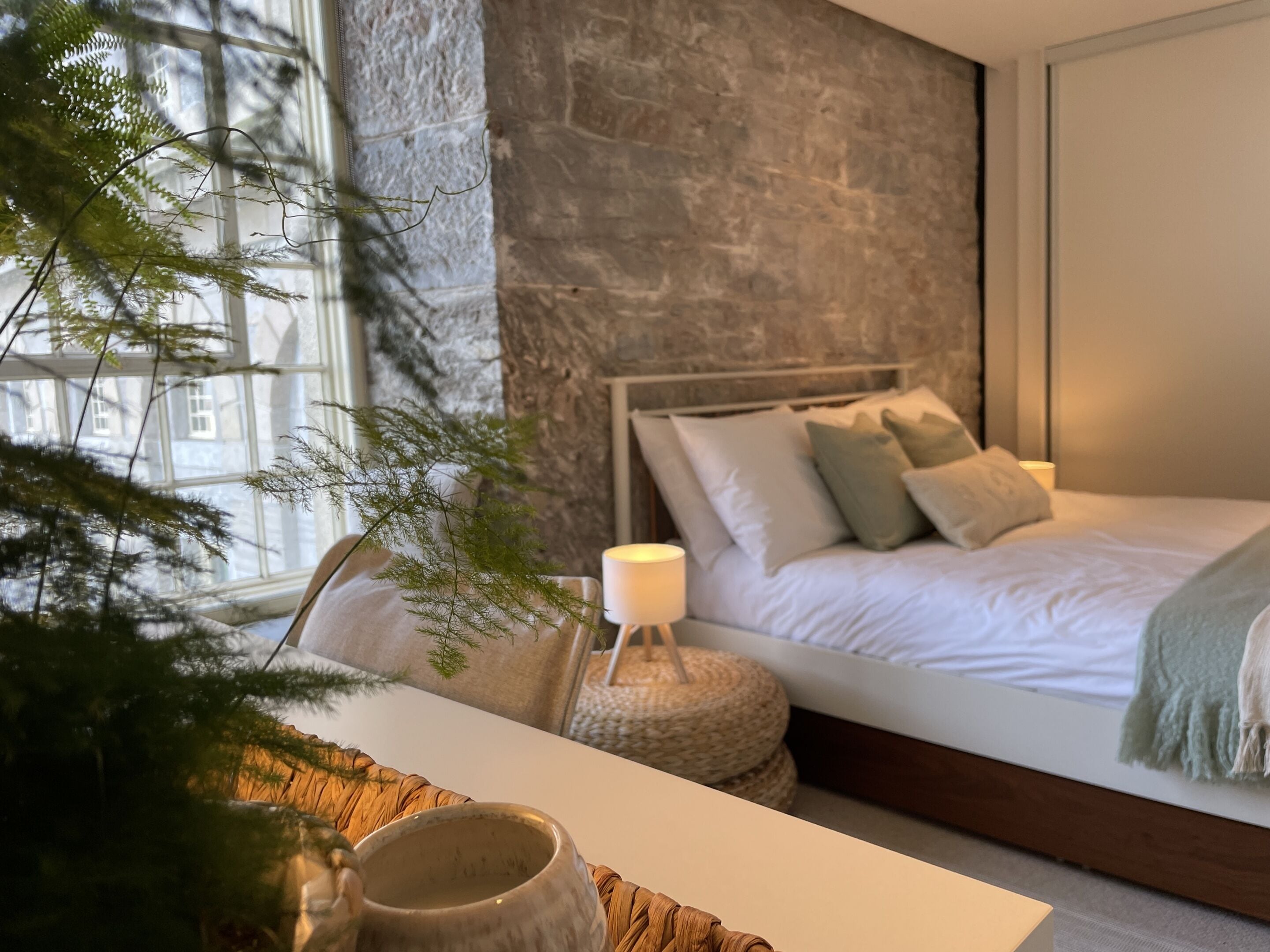 A bedroom with a stone wall, a bed with pillows, a bedside lamp, and a window with a view of buildings.