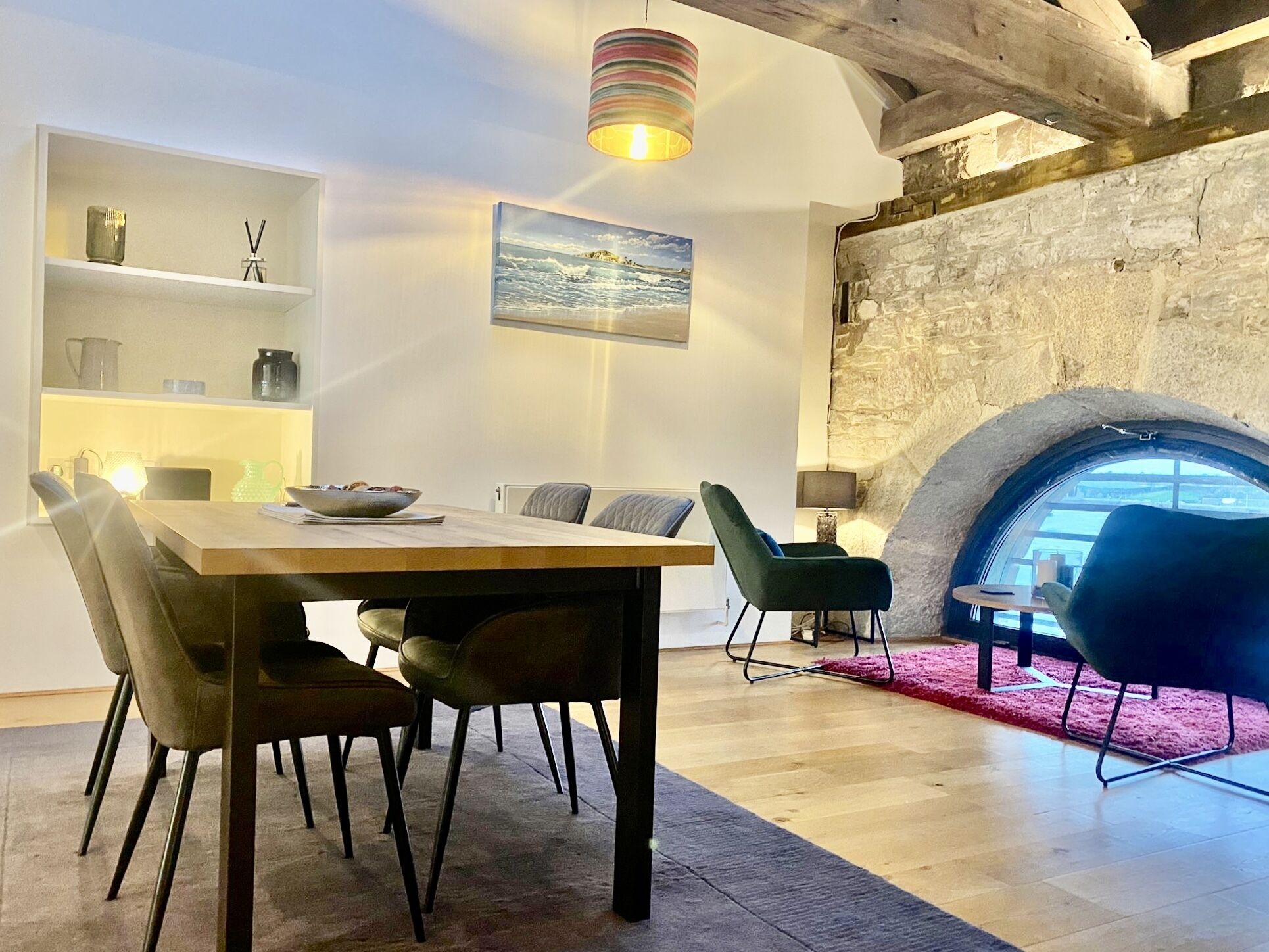 A modern dining area with a wooden table and chairs, a stone wall with a circular window, and a hanging lamp.