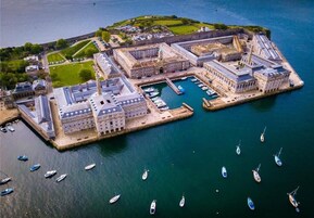 An aerial view of a historic port with a large building complex, a marina with numerous boats, and a small island with greenery and buildings.
