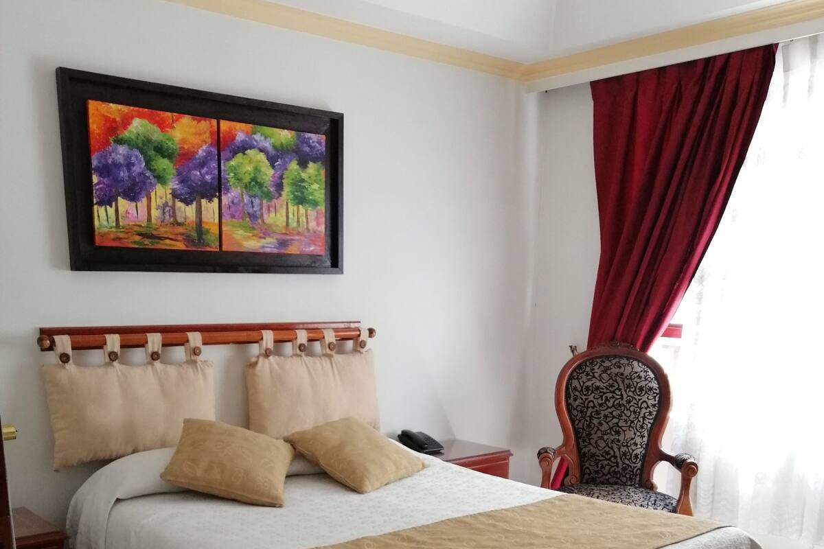 A neatly arranged bedroom with a bed, a chair, a painting, and a window with curtains.