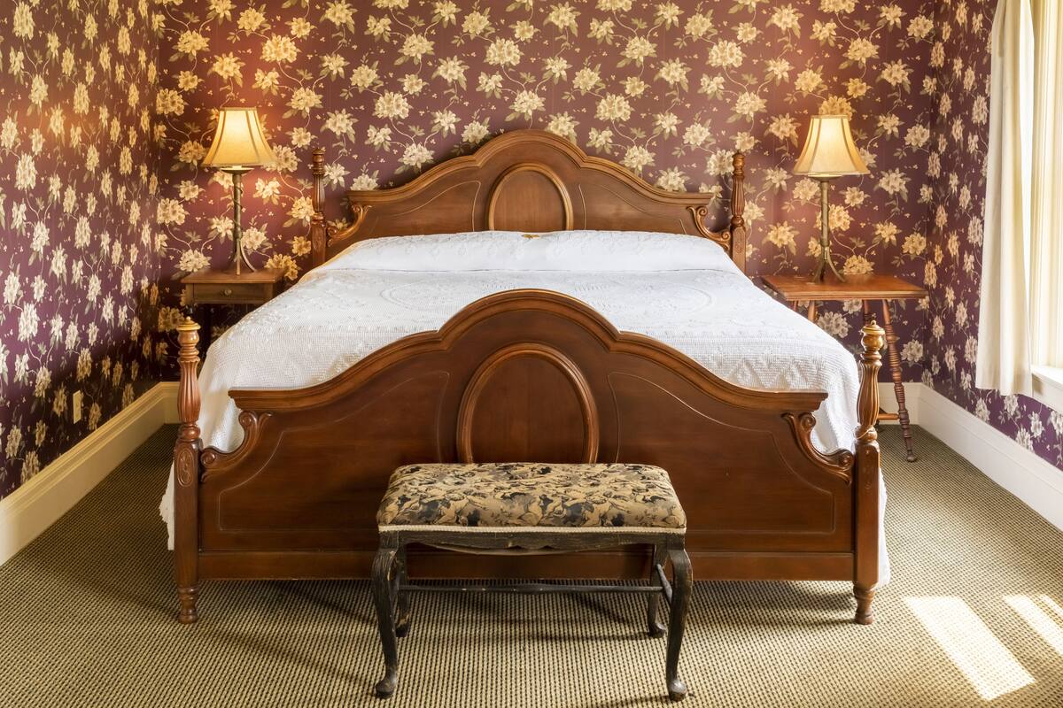 Ein Schlafzimmer mit einem hölzernen Bett, zwei Nachttischen mit Lampen, einer Bank und floraler Tapete.