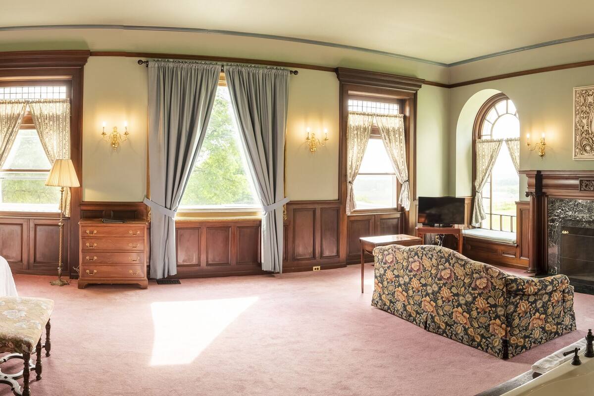 Ein geräumiges Schlafzimmer mit einem großen Bett, einer Sitzecke, einem Kamin und mehreren Fenstern mit Vorhängen.
