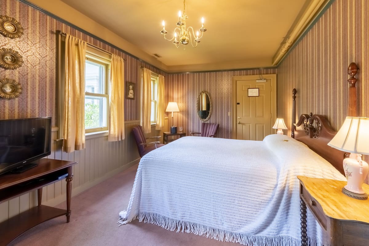 Ein Schlafzimmer mit einem großen Bett, einem Fernseher auf einem Holzständer, einer Chandelüster und rautenförmigem Wandteppich.