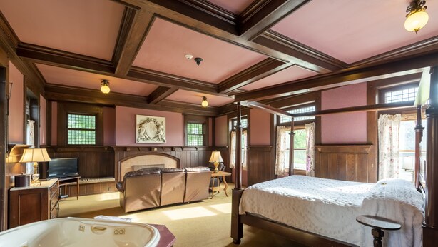A spacious bedroom with a large bed, a sitting area with a sofa and armchair, a fireplace, a large window with curtains, and a television on a wooden cabinet. - White Springs Manor a Belhurst Property (Geneva)