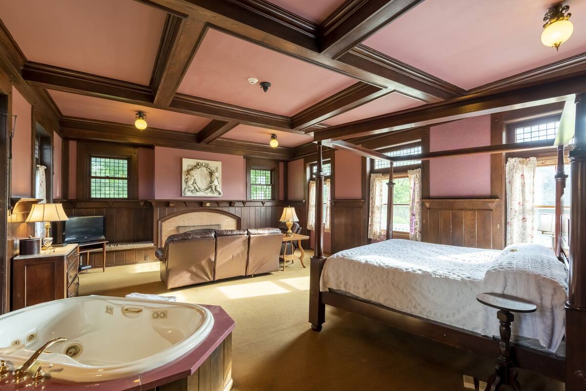 Ein geräumiges Schlafzimmer mit einem großen Bett, einer Sitzecke mit Sofa und Sesseln, einem Kamin, einem großen Fenster mit Vorhängen und einem Fernseher auf einem Holzschrank.
