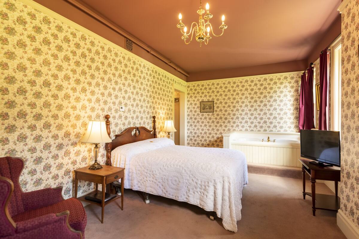 Ein Hotelzimmer mit floraler Tapete, einem Bett mit weißer Bettdecke, einem Nachttisch aus Holz mit Lampe, einem roten Sessel, einem Fernseher auf einem Beistelltisch und einer Badewanne.
