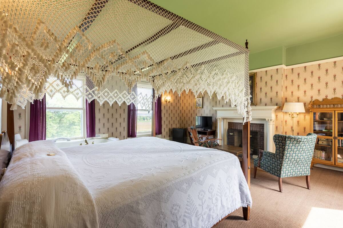 Ein geräumiges Schlafzimmer mit einem großen Bett, einem Himmelbett, einem Fernseher, einem Kamin und einem grün gemusterten Sessel.