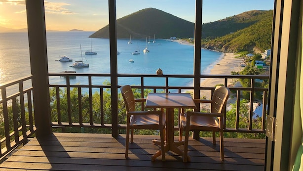Eine Holzterrasse mit Tisch und Stühlen, mit Blick auf eine Bucht, in der Boote und ein Strand liegen.
