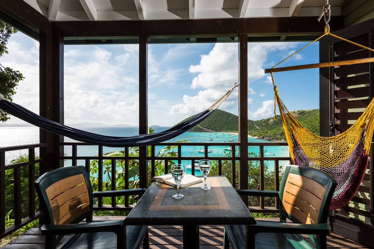 A wooden deck with a table and chairs, a hammock, and a view of a coastal landscape with mountains and turquoise waters.