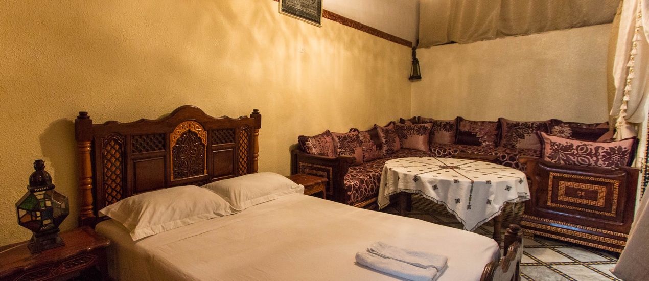 A room with a wooden bed, a small table with a tablecloth, and a sofa with patterned cushions.
