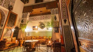 An interior with a high ceiling, ornate wooden beams, a patterned tile wall, and a seating area with a table and chairs. - Riad Meski (Fes)