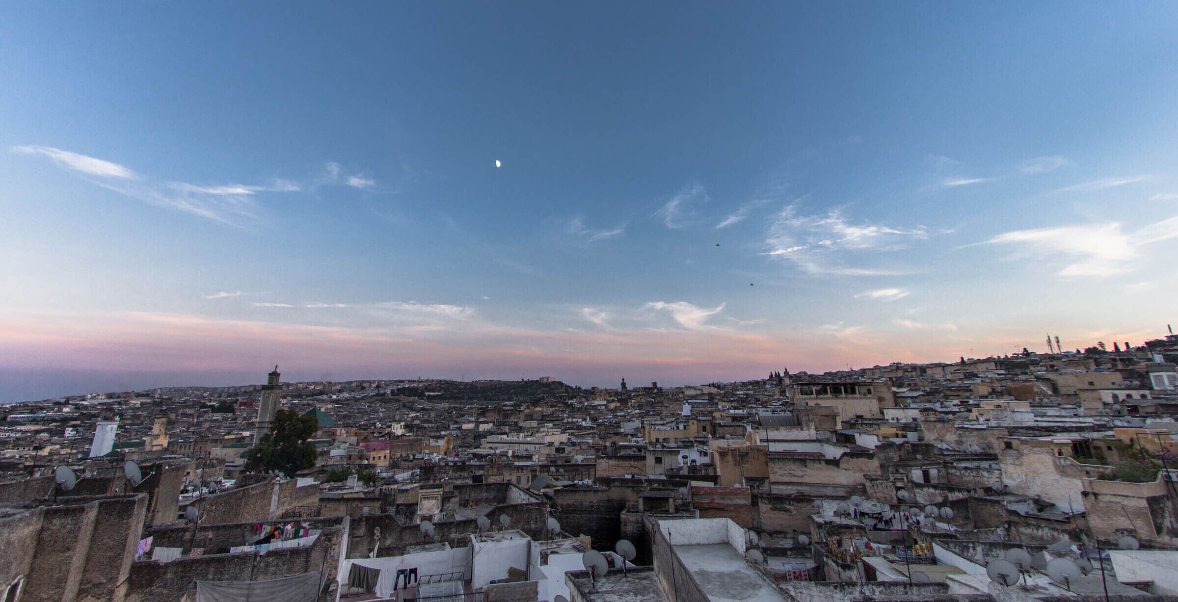 A densely packed urban area with numerous rooftops and buildings, under a clear sky with a few clouds.