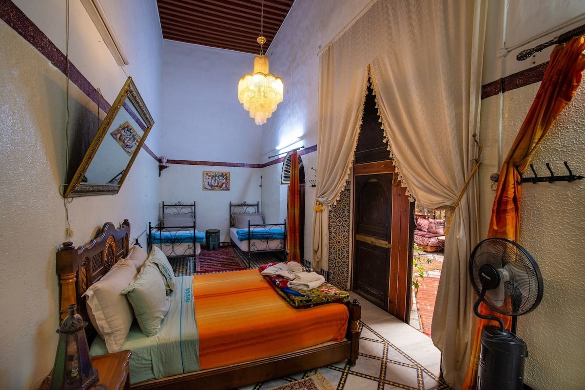 Une chambre marocaine traditionnelle avec un grand lit, deux petits lits, un lustre, un ventilateur et une vue sur la cour.