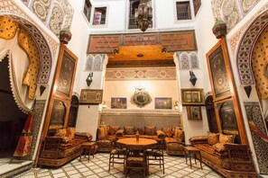 A traditional Moroccan interior with ornate arches, a central seating area with cushions, and a tiled floor.