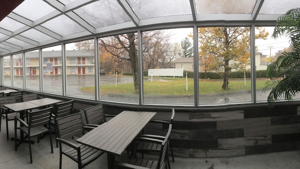 A covered outdoor seating area with tables and chairs, large windows offering views of a residential area. - Hotel Motel Hospitalité (Levis)