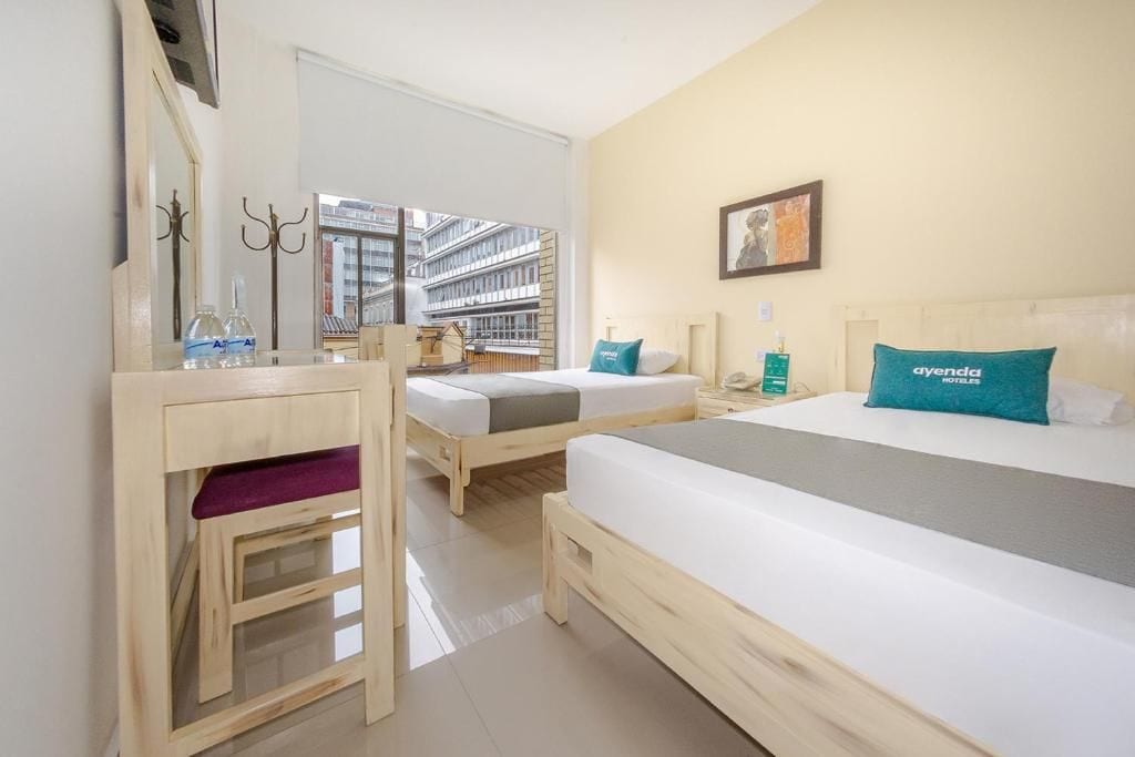 Habitación de hotel con dos camas, un escritorio de madera, un espejo y vistas a edificios.