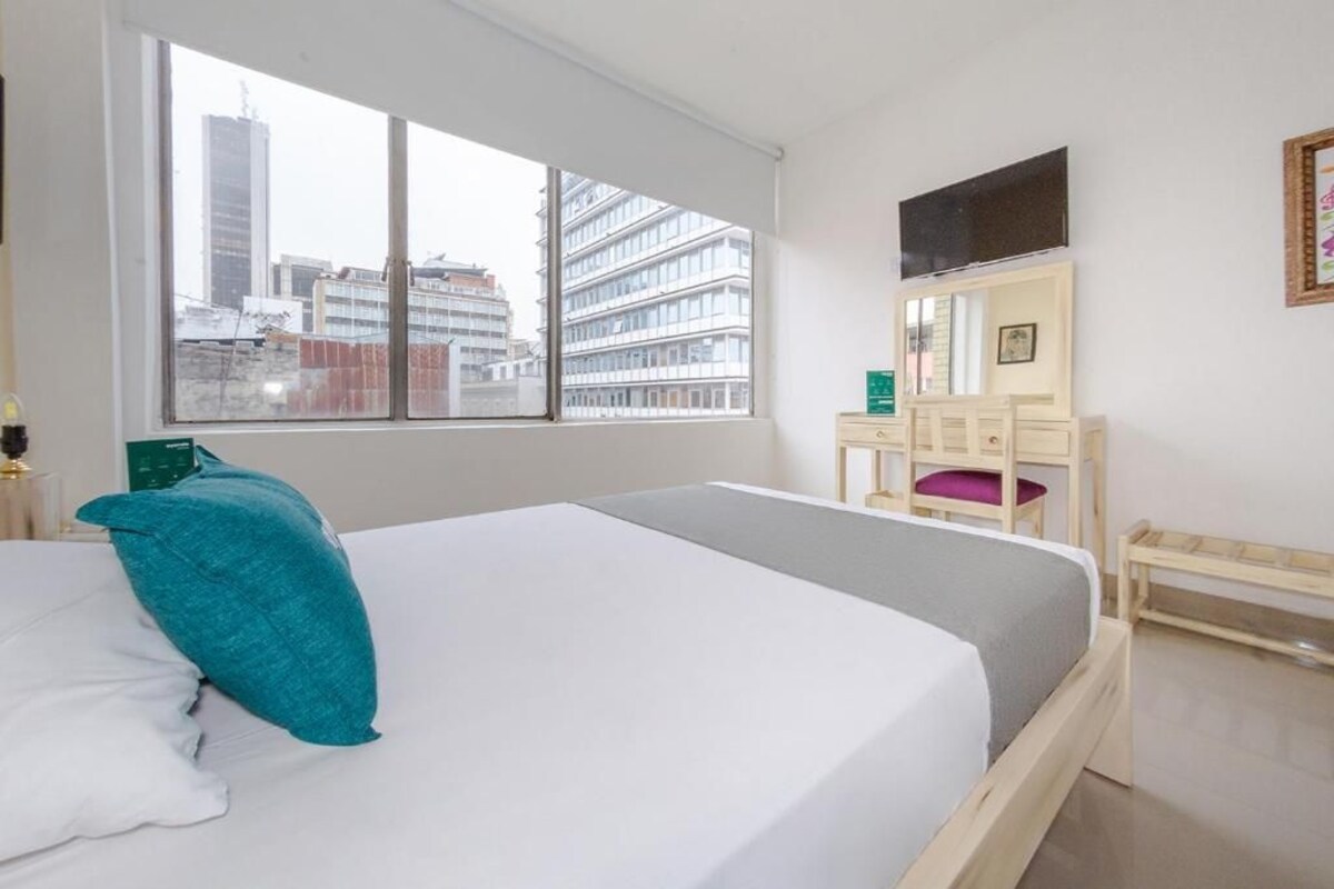 A hotel room with a large window, a bed, a desk, a chair, and a television on the wall.