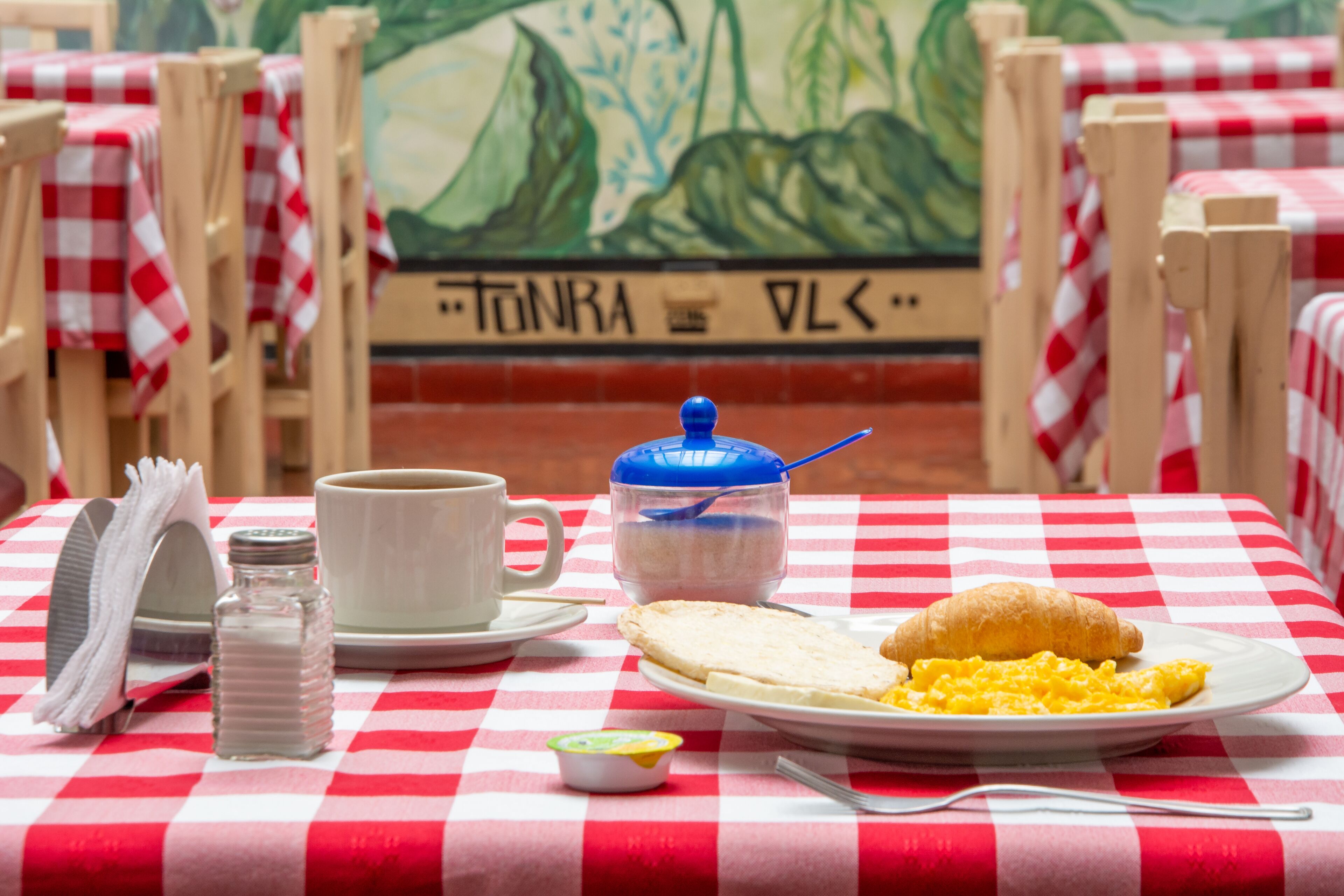 A breakfast table with a checkered tablecloth, a cup, a salt shaker, a small bowl of jam, a croissant, and scrambled eggs.