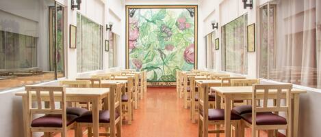 Un comedor con mesas y sillas de madera dispuestas en filas, un gran mural en la pared de fondo y ventanas amplias a los lados.