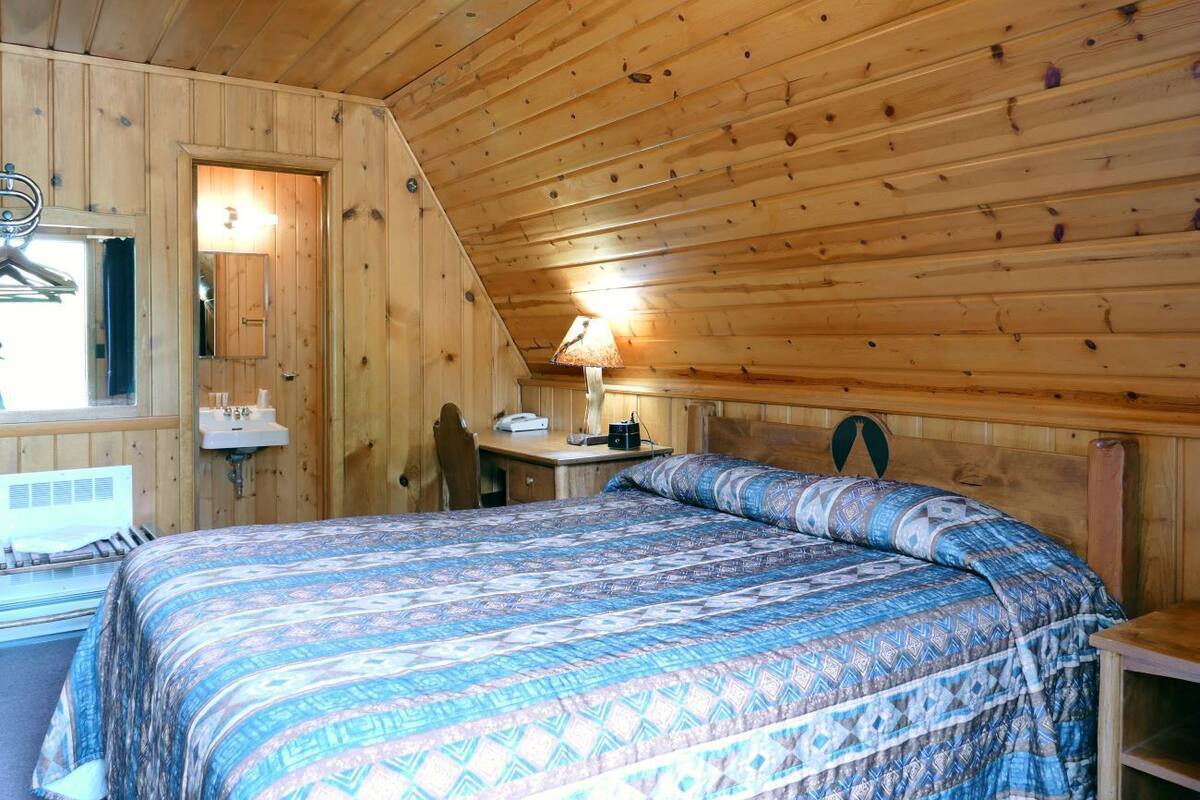 A wooden cabin room with a bed, a desk, a hanging coat rack, and a window.