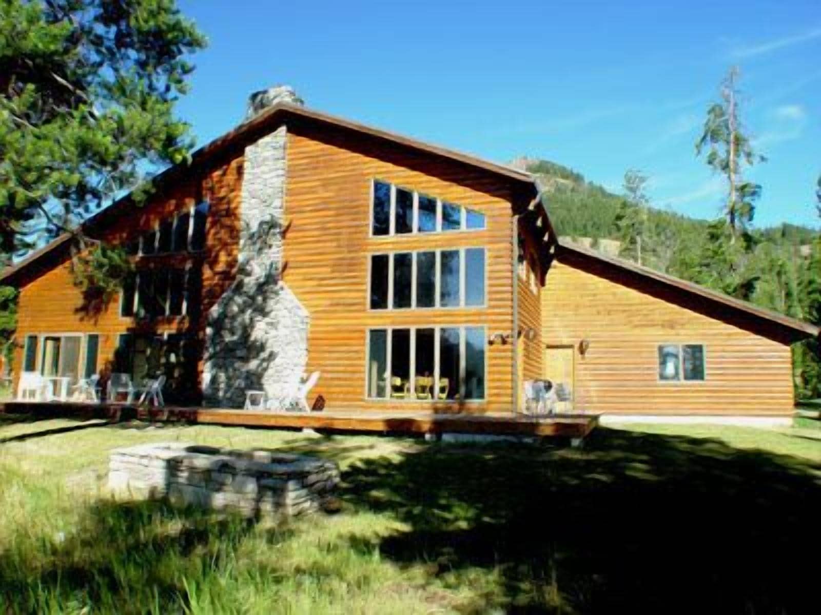 A two-story wooden cabin with a stone chimney, large windows, and a stone foundation.