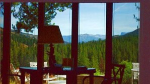 Una habitación con ventanas amplias que ofrecen vistas a un bosque y montañas. Hay mesas y sillas en el interior.
