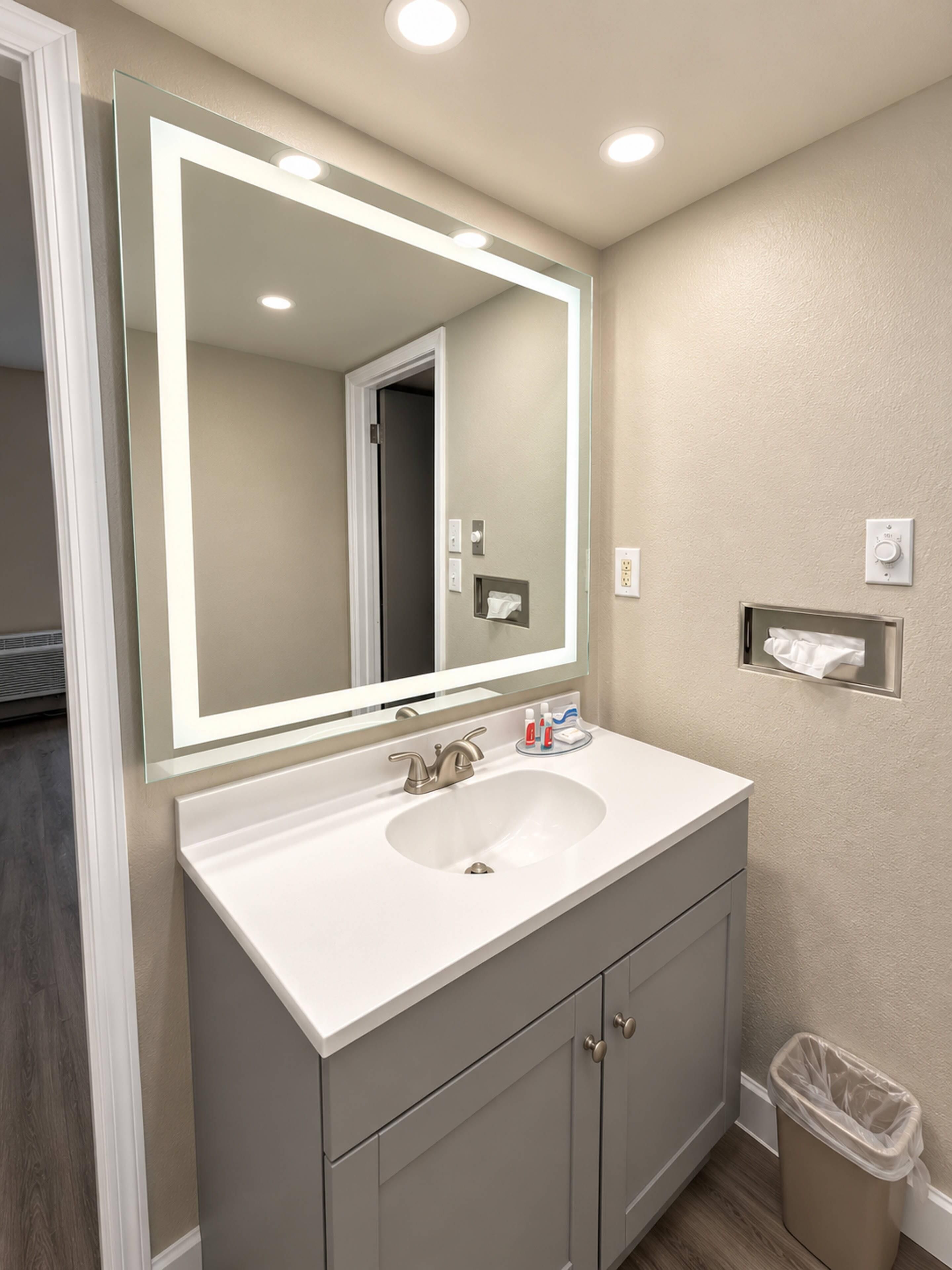 A bathroom with a large mirror, a sink, cabinets, a towel dispenser, and a trash can.