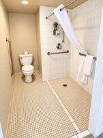 Un baño con inodoro blanco, ducha con cortina blanca y barra de apoyo.