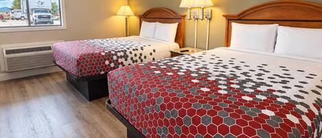 A hotel room with two beds, each with a patterned bedspread, bedside lamps, and a window with a view of parked vehicles.