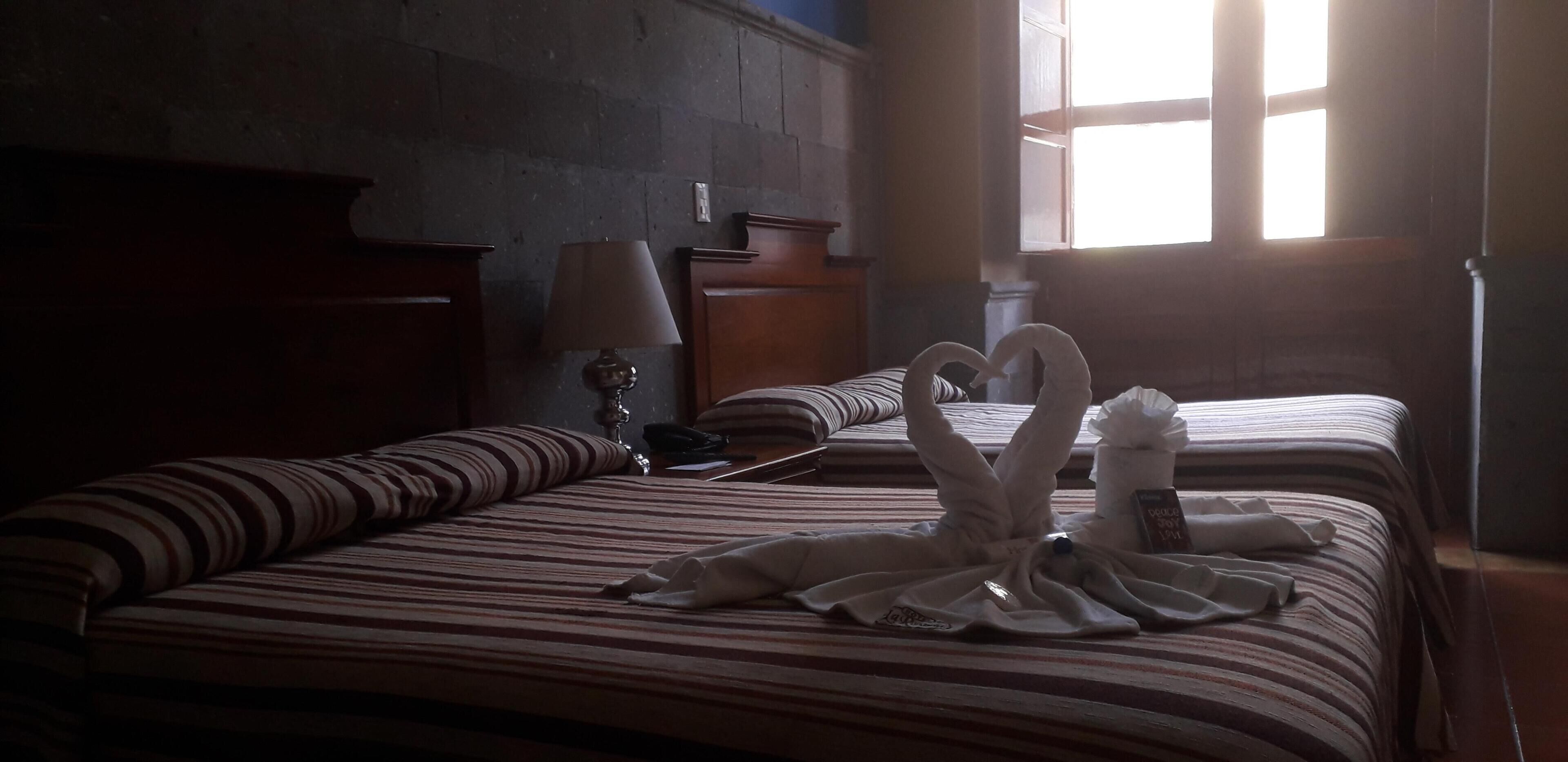 Une chambre d’hôtel avec deux lits, une literie à rayures, une lampe, une tête de lit en bois et une fenêtre laissant entrer la lumière du soleil.
