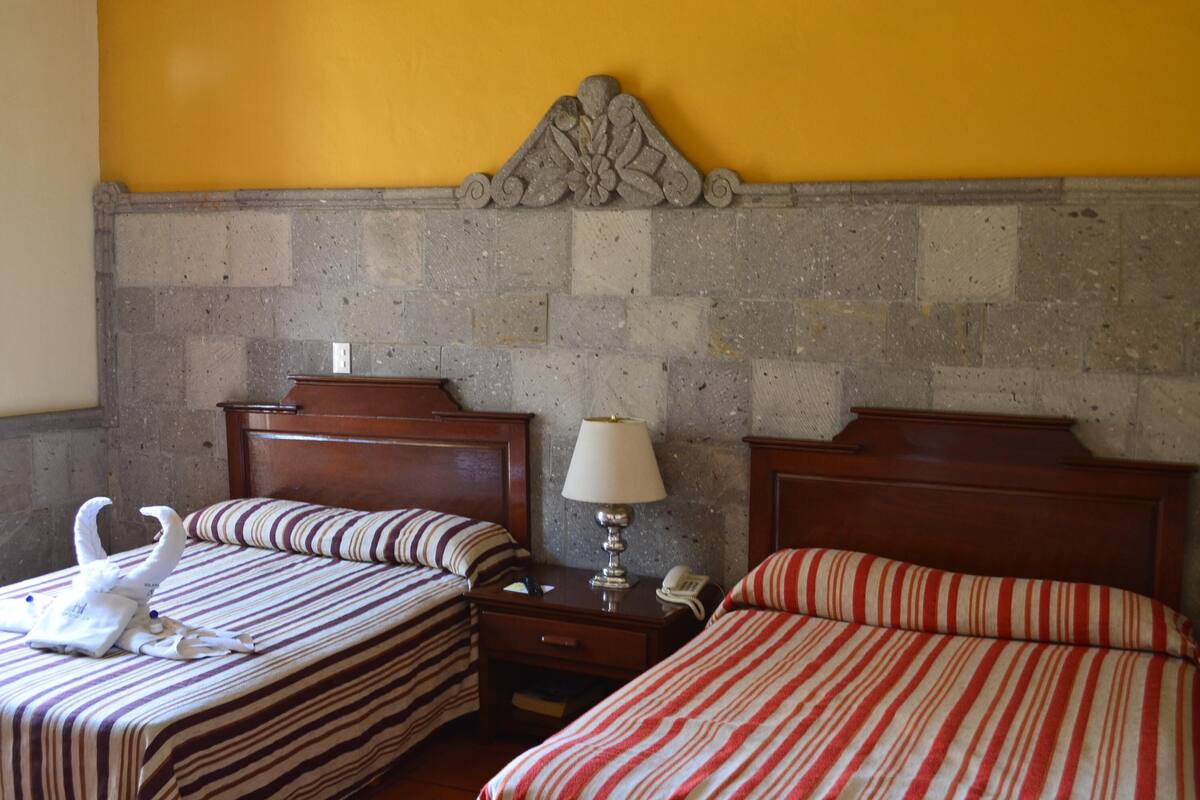 Habitación de hotel con dos camas, una mesita de noche con lámpara y una pared decorativa de piedra.