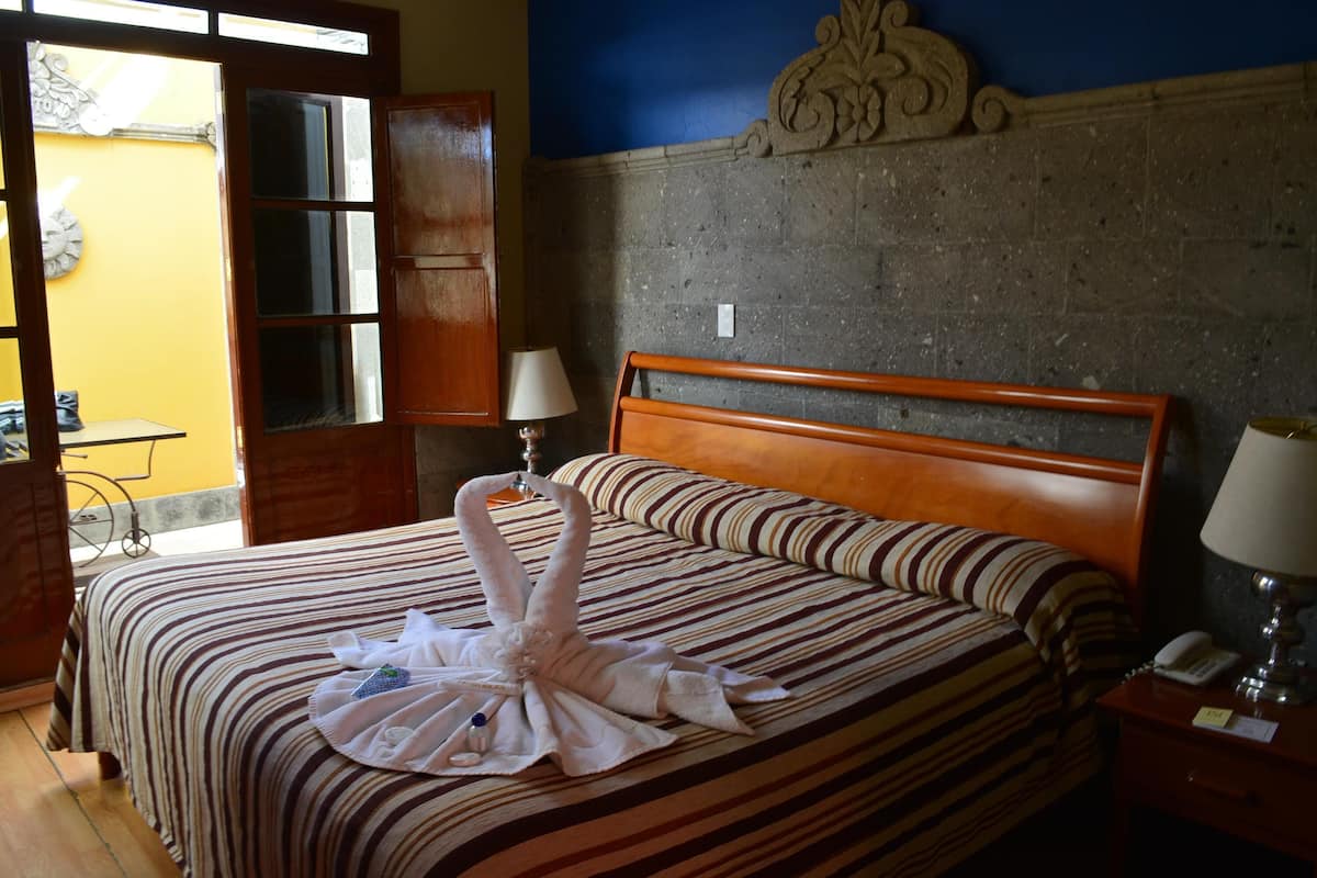 Habitación de hotel con cama, cabecera de madera, mesita de noche con lámpara y ventana con vistas al exterior.
