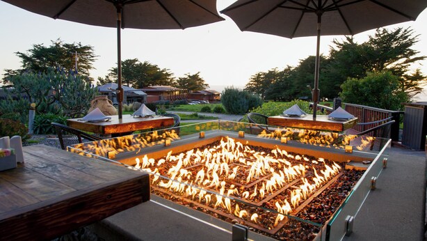 An outdoor patio area with a central fire feature surrounded by tables and chairs, umbrellas, and a view of the surrounding landscape.