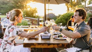 A couple toasting with wine glasses at an outdoor dining table with umbrellas, surrounded by greenery.