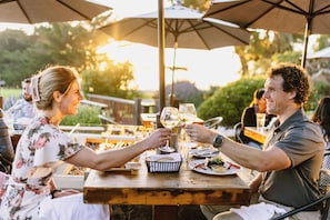 A couple toasting with wine glasses at an outdoor dining table with umbrellas, surrounded by greenery.