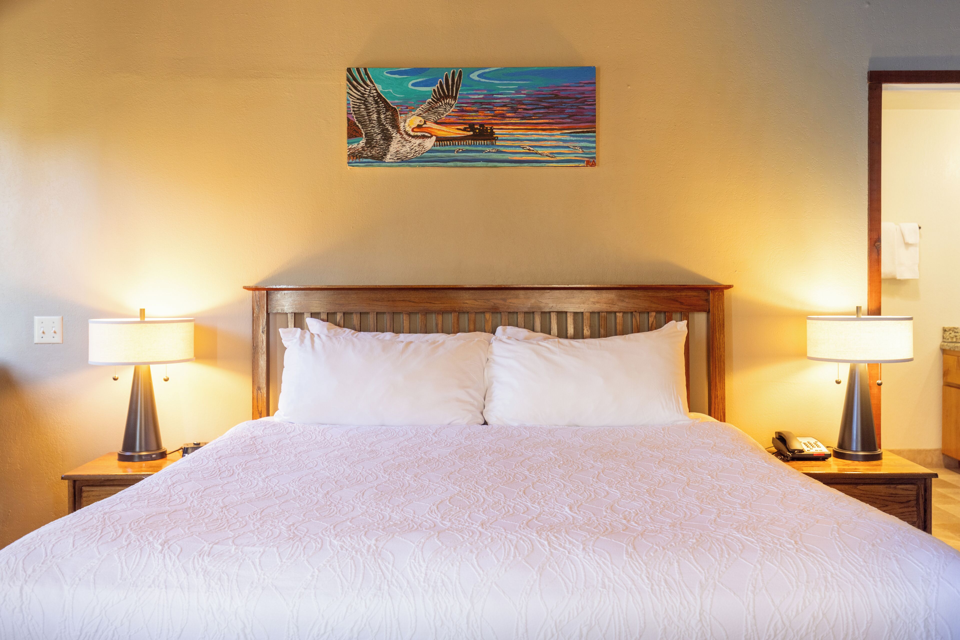 A hotel room with a large bed, two bedside tables with lamps, a painting on the wall, and a wooden headboard.