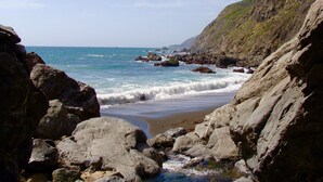 A rocky coastal landscape with waves crashing against the shore, a clear sky, and a distant view of the ocean.
