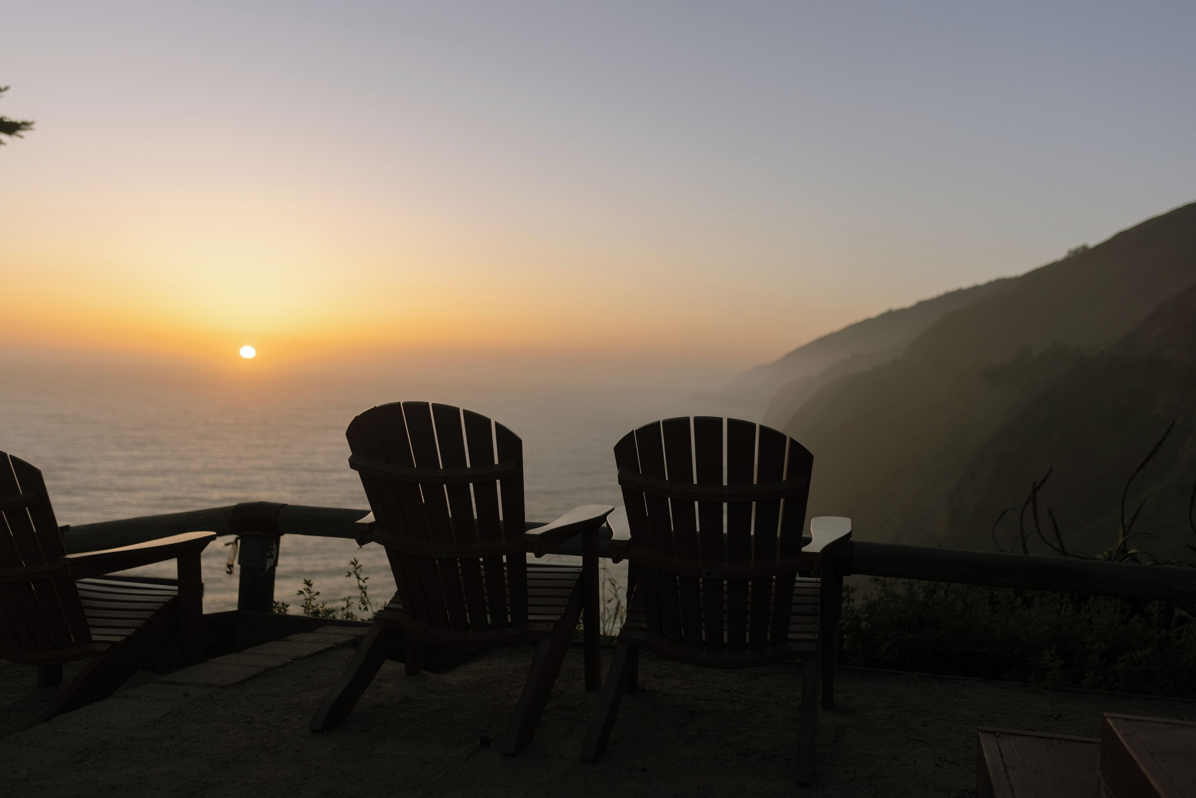 A sunset view from a beachside seating area with Adirondack chairs and a table.