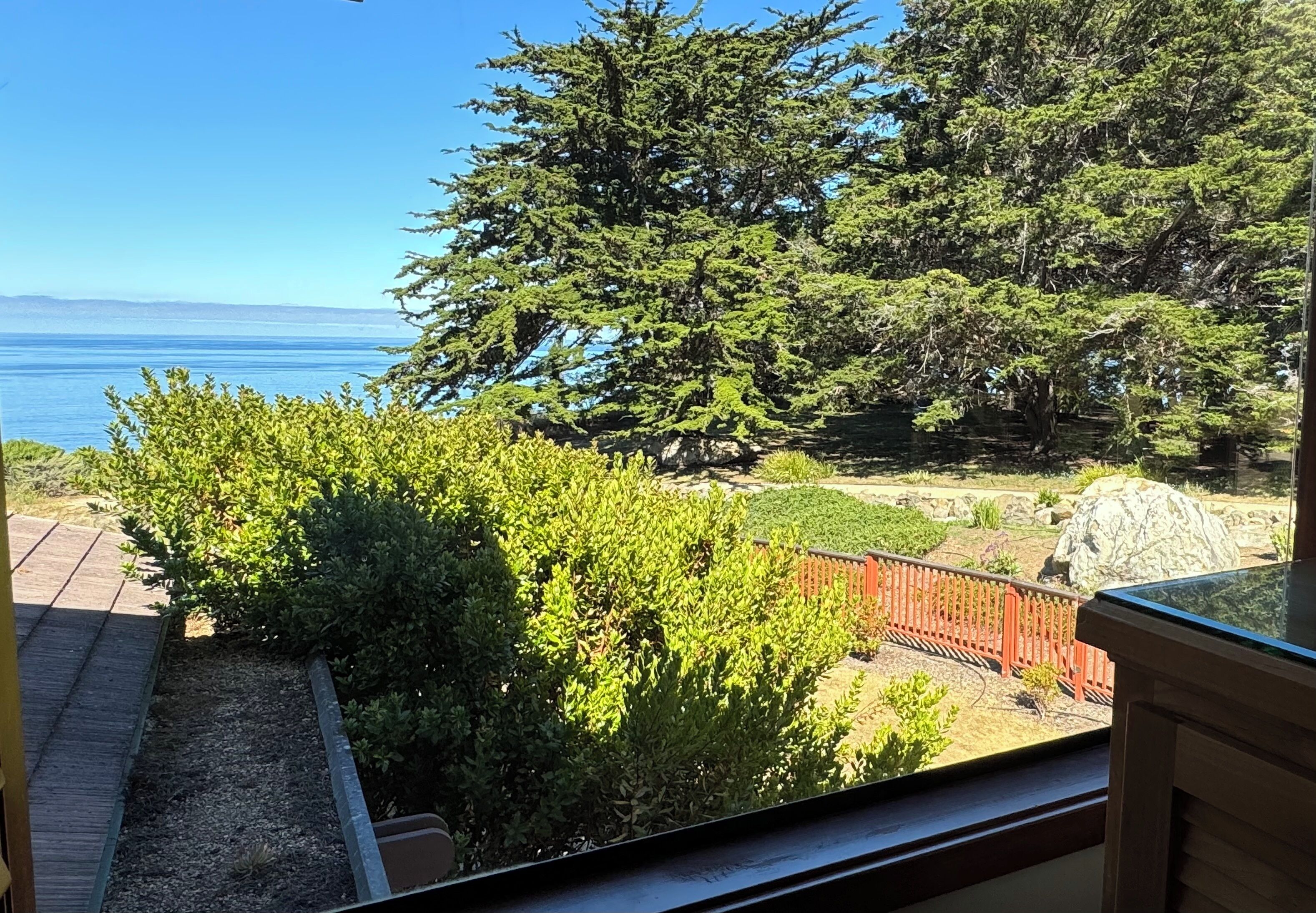 Vista de uma varanda com vista para um jardim, que inclui um deck de madeira, uma cerca vermelha e uma grande árvore.