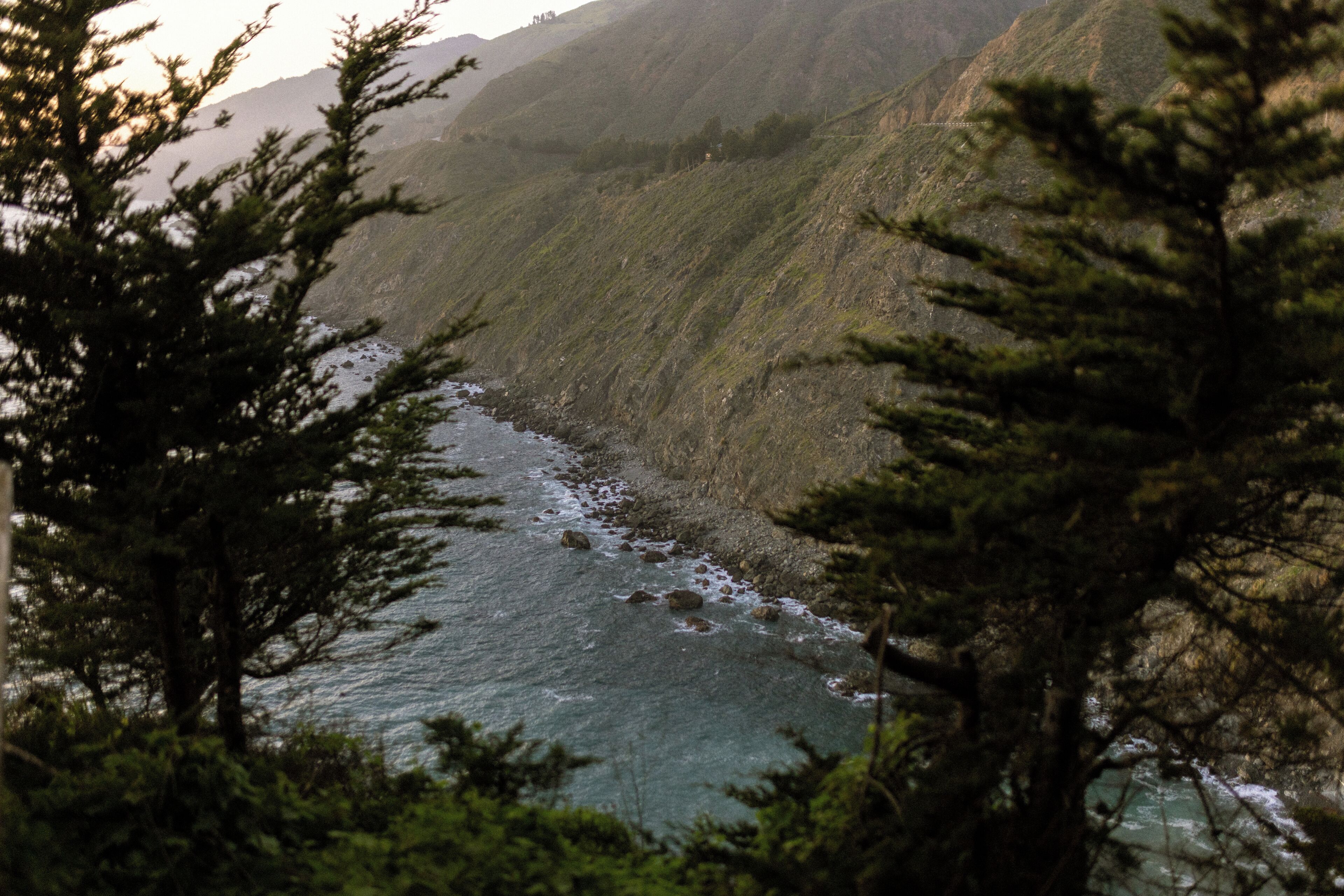 A coastal landscape with a river flowing through a mountainous area, surrounded by dense greenery.