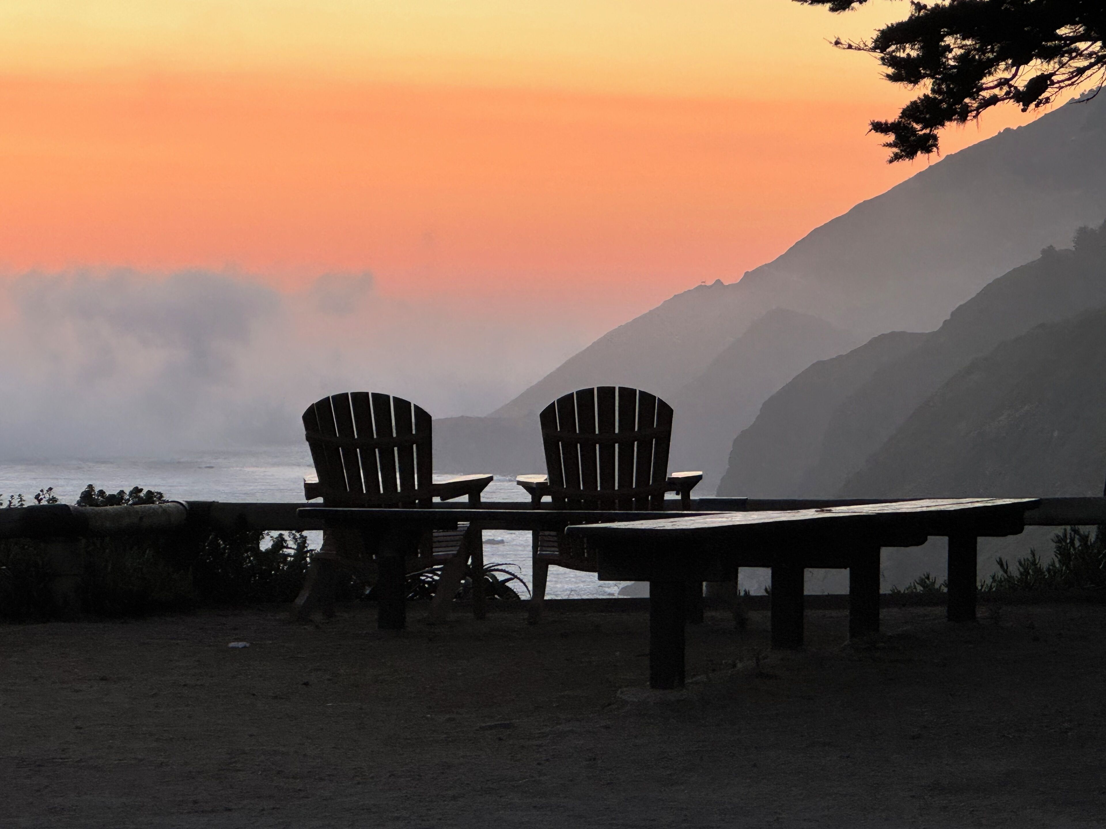 Dos sillas Adirondack y una mesa de madera sobre una playa arenosa, con vista al océano y montañas al fondo.