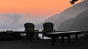 Dos sillas Adirondack y una mesa de madera sobre una playa arenosa, con vista al océano y montañas al fondo.